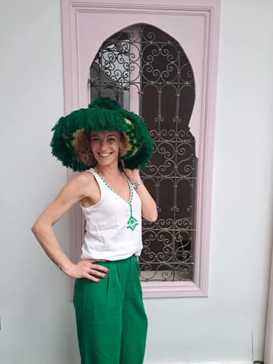 Femme souriante en tenue verte et chapelier large, posant devant une fenêtre décorative.